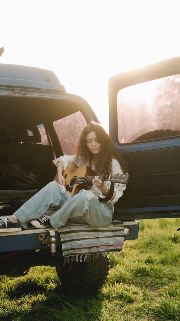 Women in the 70's era sitting in the back of her van planning the guitar in the sunshine outdoors. This picture is representing women in the 70's time period, as we look back at the ways they are growing and advancing in the workplace and in their careers, and how they can still continue to grow and advance.